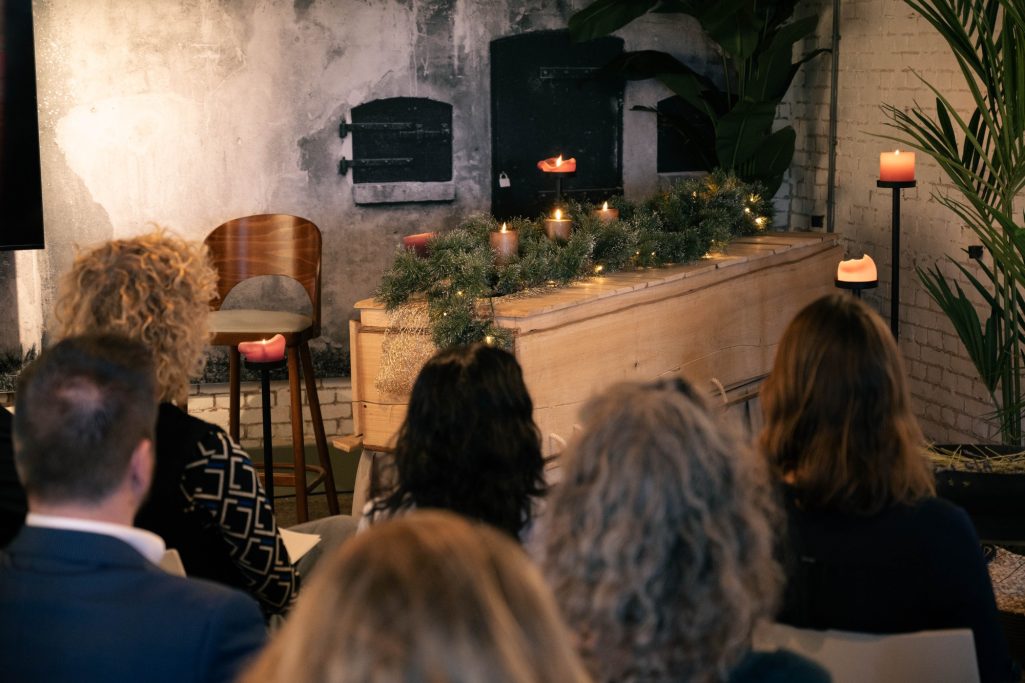 Een zaal met een eenvoudige kist, omringd door kaarsen en planten, tijdens een ceremonie.